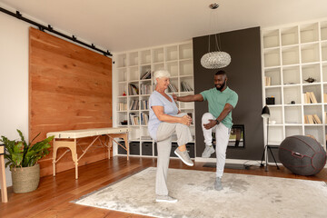 African american male physiotherapist helping caucasian senior woman to stretch her leg at home