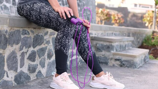 Young woman sitting with a skipping rope. Doing sport outside. 4K