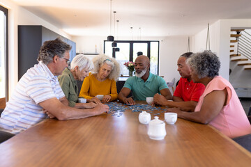 Multiracial senior friends playing jigsaw puzzle on wooden table while sitting in nursing home