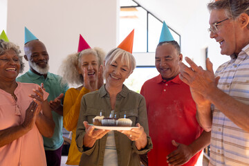 Multiracial friends clapping and singing for happy senior woman holding birthday cupcakes