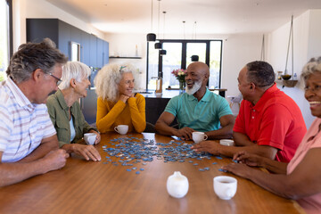 Multiracial seniors friends playing jigsaw puzzle while sitting at wooden table in nursing home