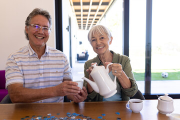 Portrait of smiling asian senior woman pouring coffee in cup held by caucasian senior man at table - Powered by Adobe