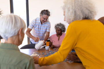 Caucasian senior man serving coffee to multiracial friends playing jigsaw puzzle in nursing home