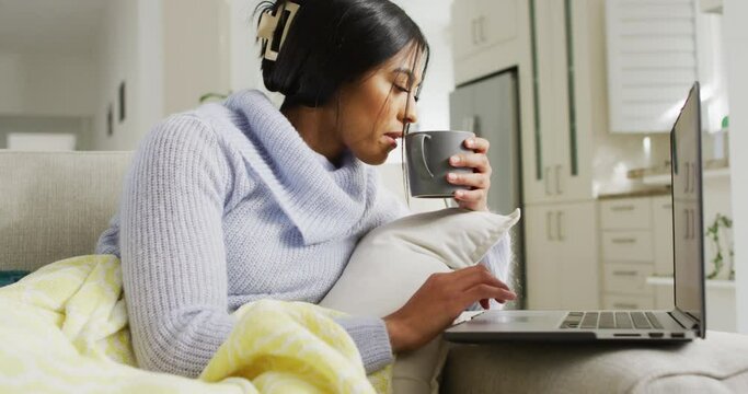 Video of happy biracial woman in hijab sitting on sofa with coffee and using laptop