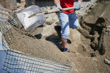 Close up of man with shovel in his hands throwing cementitious floor screed material into container with net. Male in work gloves shoveling sand-cement mix outdoors at construction site.