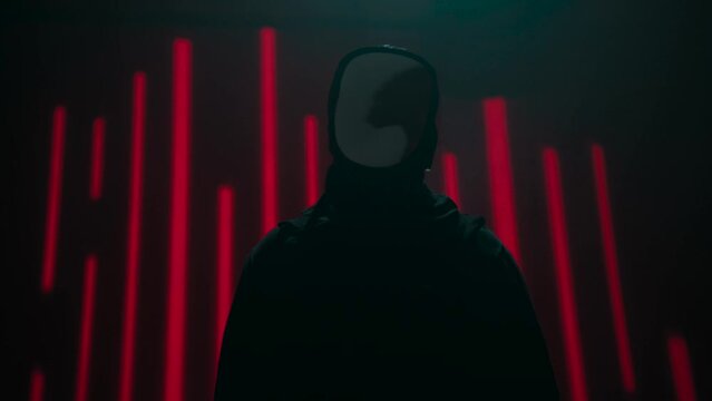 An anonymous hacker is standing in the dark in front of red bars. The silhouette looks like its speaking.