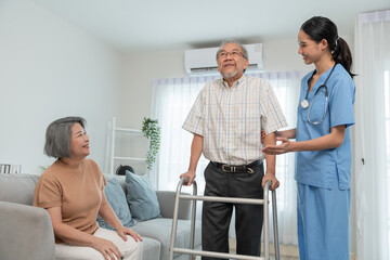 Asian young woman caregiver doctor during home visit helping elderly man patient to walk using walker frame while older woman his wife sitting on sofa encourage to him