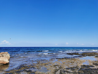 The coast of the Mediterranean Sea, waves, clear water, a stone ridge against a blue sky with clouds.