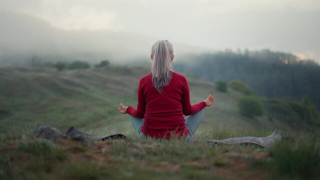 Rear view of senior woman doing breathing exercise in nature on early morning with fog and mountains in background.