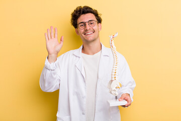 young handsome guy smiling happily, waving hand, welcoming and greeting you. spine specialist...