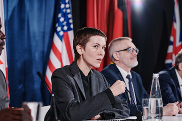 Fototapeta na wymiar Young female speaker speaking to microphone answering questions during press conference with other colleagues in background