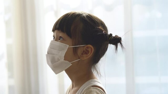Asian father putting on medical disposable mask on his cute daughter face in living room. Father and kid going out during COVID-19 pandemic, parent helping little girl to wear protective face mask.