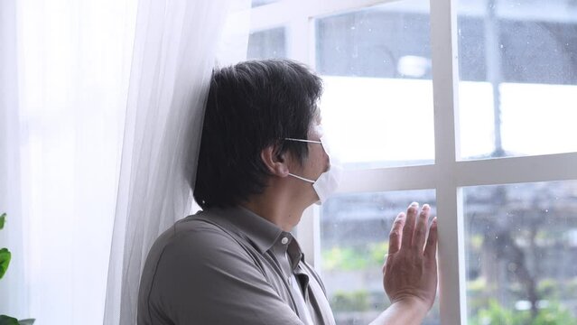 Sad Middle-age Asian man with medical mask looking out the window being quarantined at home. Quarantine concept during the coronavirus pandemic.
