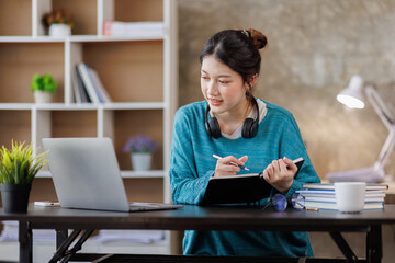 Young attractive female student writer scribing notes to a textbook to prepare for the  Exam preparation, Asian girl sitting homework on a laptop computer, making a plan for tomorrow creating a list