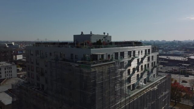 Fly over apartment building with rooftop terraces and scaffolding around facade. Revealing multilane expressway and panoramic view of industrial or logistic site. New York City, USA
