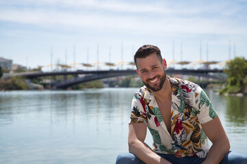 Young and handsome man, blue eyes, perfect smile, beard and Hawaiian shirt, sitting on a pier by...