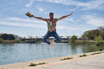 Young and handsome man, beard, hat, Hawaiian shirt and jeans, jumping in the air happily on a pier...