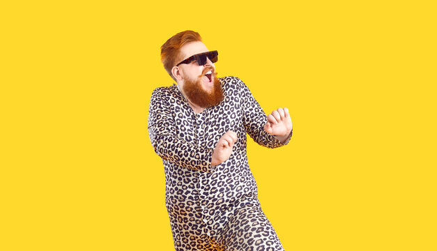 Crazy Plus Size Man In Funny Pajamas Dancing And Having Fun In Fashion Studio. Happy Bearded Fat Guy Wearing Cool Sunglasses And Comfy Leopard PJs Singing And Dancing Isolated On Yellow Background