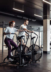Obraz na płótnie Canvas Athletic man and woman doing intense workout together by pedaling air bike in modern gym. Healthy lifestyle