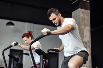 Athletic man and woman doing intense workout together on air bike in modern gym. Healthy lifestyle
