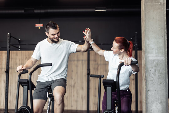 Smiling man and woman doing intensive workout together by pedaling air bike and high five in modern gym. Healthy lifestyle - Powered by Adobe