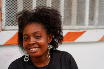 A smiling Haitian woman with a bouffant hairstyle sits outside her house.