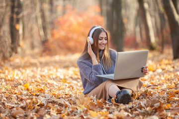 Young woman in headphones with a laptop sits on fallen leaves in autumn forest and has fun listening to music