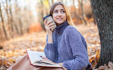 Young woman sitting on fallen leaves with book and coffee cup in autumn forest