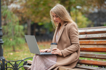 A young business woman in a coat works with a laptop while sitting on a bench in a city park. Freelance, remote work