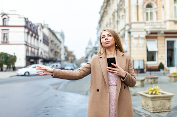 Obraz premium Blonde woman catching car, holding smartphone with hand gesture in european city