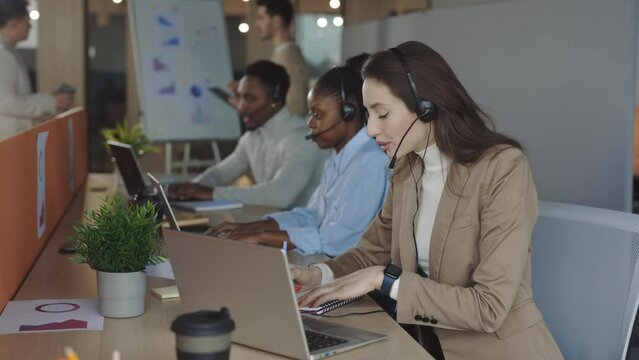 Concentrated woman operator in headset talking with client on videoconference and writing remarks on notebook. Diverse teammates of marketing group using modern laptops in office.