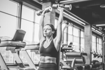 Fototapeta na wymiar Fit woman trains shoulders with dumbbells in her hands doing dumbbell bench press over her head while sitting on a bench in the gym