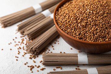 Buckwheat porridge in a bowl and buckwheat soba noodles on a white marble background. Raw buckwheat. Ingredients for gluten free cereal. Healthy food. Place to copy.