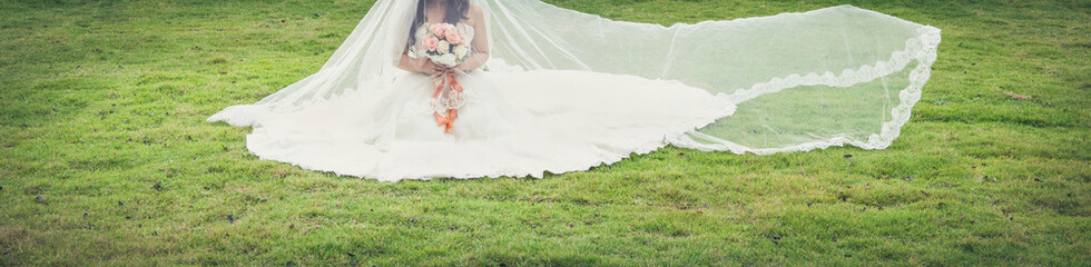 Fototapeta na wymiar Bride with a bouquet in her hands sitting on the green grass in the park. Concept of bride banner size