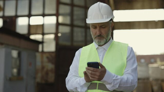 Focused mature foreman in reflective vest and white helmet using cell phone while standing at construction site. Thoughtful man checking building information on modern smartphone.