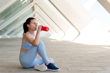 Fototapeta na wymiar Fit, sporty woman sitting drinking water from a glass bottle after exercise or running. Urban background.