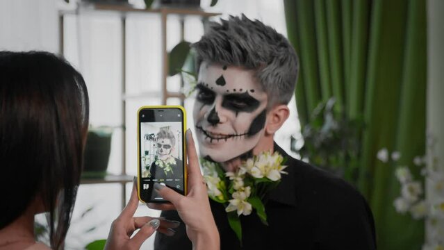 Female hands take a picture on the phone of a guy in a creepy image for Halloween. A young man prepares makeup for Halloween. Create a Halloween look
