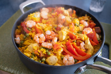Shallow focus on prawns in a paella dish resting after cooking. There is steam from the heat. They are in a large frying pan with chorizo sausage, rice and chicken with onion, peas and peppers.