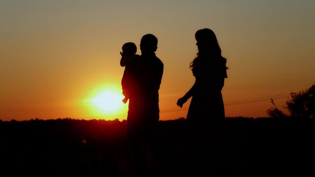 Caring father holds the baby on his arms, mom comes up and hugs the family, unrecognizable silhouette at sunset, paternal love