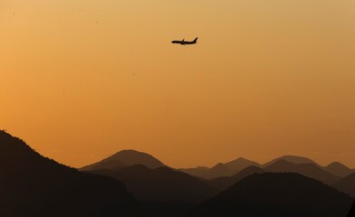 Jet plane flying over the mountains at sunrise. Silhouettes and various shades of yellow and orange.