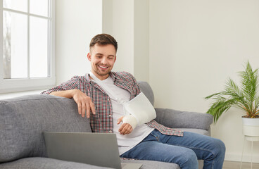 Happy young Caucasian man with broken arm in cast spending rehabilitation time at home, sitting on comfortable sofa in living room, watching video on laptop computer, enjoying good movie and smiling