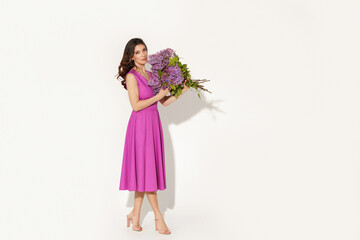 Full-length studio fashion portrait of elegant woman wearing purple dress and holding lilac brunches against white wall. International woman’s day, Mother’s day and spring concept.