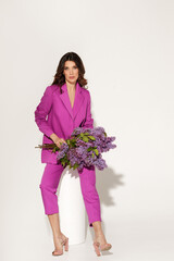 Full-length studio fashion portrait of elegant woman wearing purple suit and holding Lilac brunches against white wall. International woman’s day, Mother’s day and spring concept.