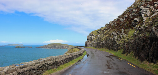 Road between grassy stone slope and sea in western Ireland in blue overcast weather still wet and full of puddles from the rain