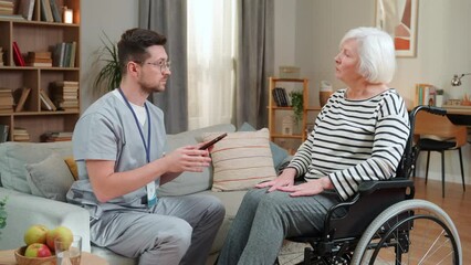 Handicapped senior Caucasian woman in wheelchair talking with young handsome male nurse, man typing on tablet device. Retirement, healthcare nursing, health support, social assistance, doctor concept