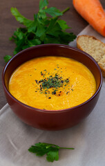 bowl of tasty carrot cream soup with fresh carrots and herbs on wooden background. Carrot soup with parsley on brown table. Spring vegetable soup. Homemade cream soup with copy space. vertical