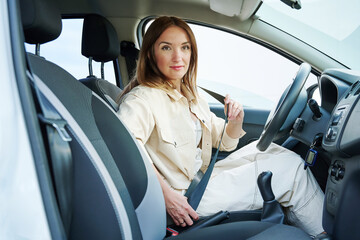 Young happy woman sits at the wheel of a car and hold seat belt looks at window. Side view