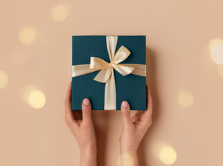 Female hands with natural manicure holding blue gift box with light golden ribbon on trendy beige...