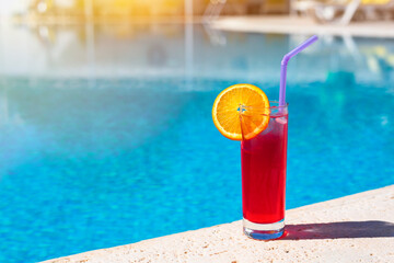 A red cocktail in a glass with an orange slice on the edge of the hotel pool. Bright sunlight, close-up, copy space.