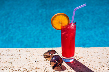 Red cocktail in a glass with an orange slice on the edge of the pool. Refreshing iced drink on a hot sunny day.